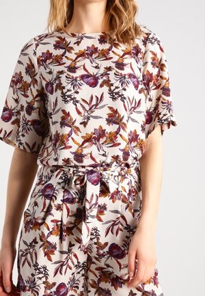 Kvinde iført en hvid kortærmet jumpsuit med et lilla og orange blomsterprint og et bundet stofbælte i taljen.