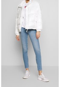 Chaqueta tipo puffer blanca con cuello alto, de textura suave y cierre de cremallera, llevada sobre una camiseta blanca con un logo rojo y unos vaqueros azules.