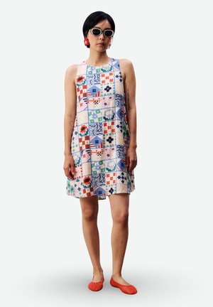 Femme portant une robe sans manches patchwork imprimé floral, lunettes de soleil blanches, boucles d’oreilles fleurs rouges et chaussures rouges sans lacets, debout sur fond blanc.