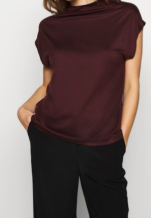 Kvinde iført en kortærmet bordeaux top og sorte bukser med hænderne i lommerne, set fra skuldrene til hofterne mod en ensfarvet baggrund.