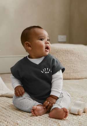 Bebelușul poartă un tricou „Little Man”, stând desculț pe un covor texturat, uitându-se înspre lateral cu gura deschisă lângă o mașinuță de lemn.