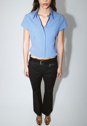 Frau trägt eine kurzärmelige blau karierte Bluse, schwarze Hose mit Gürtel und goldene Sandalen, steht vor hellem, einfarbigem Hintergrund.