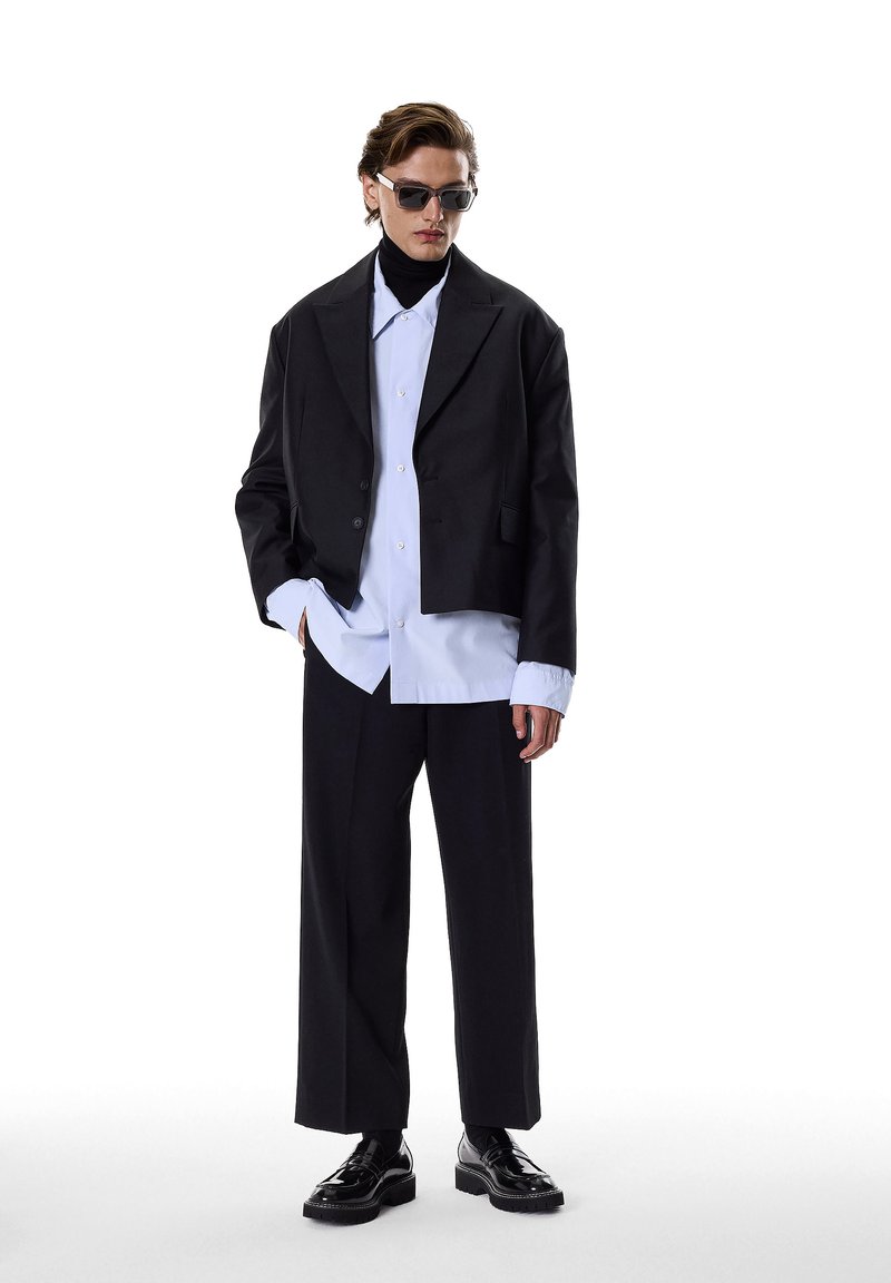 Black cropped blazer over a light blue oversized shirt, paired with black wide trousers and shiny black shoes. Accessories include sunglasses.