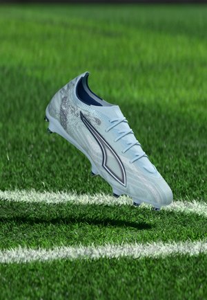 Scarpetta da calcio bianca e argento con logo nero, sospesa sopra l'erba verde vicino alle linee bianche del campo.