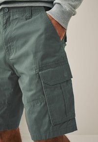 Shorts cargo en tissu vert foncé, avec poches latérales, une coupe décontractée et des détails de couture contrastés. Matériau en coton texturé.