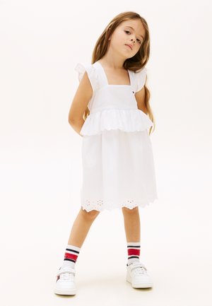 Jeune fille aux longs cheveux portant une robe blanche sans manches, des chaussettes rayées et des baskets blanches, debout les mains dans le dos.