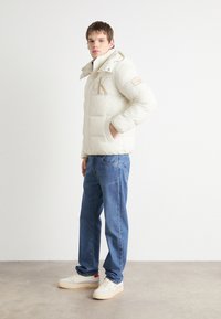 Pufferjakke i kremfarge med logo, med hette, knappeåpning og lapp på ermet. Kombinert med blå jeans og hvite joggesko.