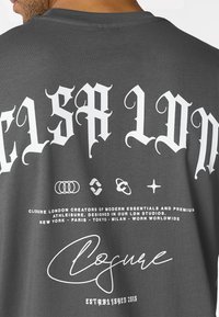 Mann trägt ein dunkelgraues Hemd mit weißem, gotischem Schriftzug "CLSPA LDN", dem Markennamen "Closure" und aufgedruckten Städtenamen auf der Rückseite.