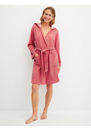 Glimlachende vrouw staat blootsvoets en draagt een knielange roze en grijs gestreepte badjas met kap en taillelint en voorzakken.