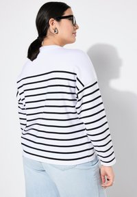 Maglione a righe bianche con linee orizzontali nere, vestibilità rilassata, scollatura rotonda e maniche lunghe, abbinato a jeans azzurri chiari.