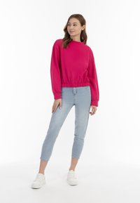 Fukszia színű, hosszú ujjú pulcsi rugalmas aljjal, világos kék slim fit farmernadrággal és fehér sportcipőkkel egy fehér háttér előtt.