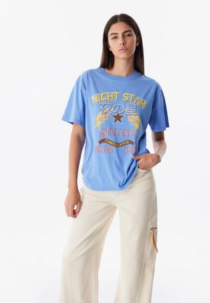 Světle modré bavlněné tričko s grafickým potiskem v žluté, červené a bílé barvě se hvězdami, spárované s krémovými kalhotami s širokými nohavicemi a bočním zipem.