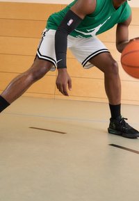 Groene mouwloze jersey met logo, zwarte armbeschermer, witte shorts met zwarte bies en zwarte basketbalschoenen met groene accenten. Basketbal in de hand.