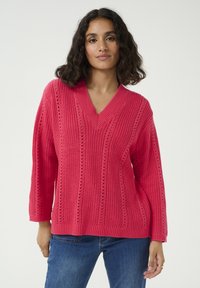 Pull en tricot rose avec un décolleté en V, présentant des motifs verticaux et des détails ajourés. Manches longues et coupe décontractée. Associé à un jean bleu.