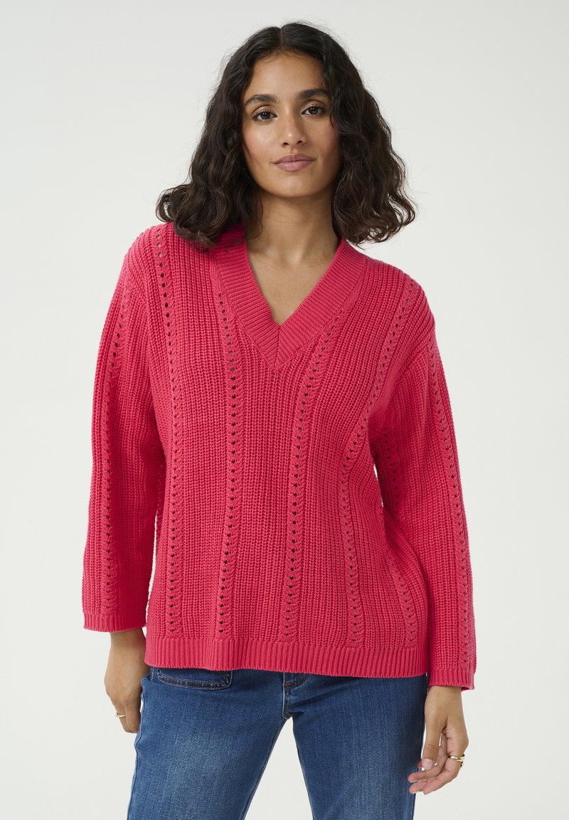 Pull en tricot rose avec un décolleté en V, présentant des motifs verticaux et des détails ajourés. Manches longues et coupe décontractée. Associé à un jean bleu.