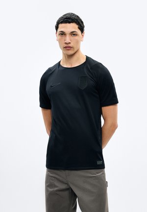 Jeune homme debout, de face, portant un maillot de sport noir Nike avec un blason discret et un pantalon gris, sur un fond clair uni.