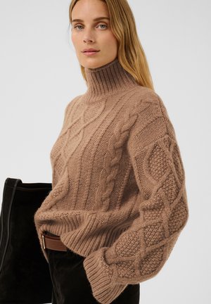 Femme portant un pull à col roulé marron en maille torsadée avec des motifs texturés et un pantalon noir, tenant un sac à main noir devant un fond uni.