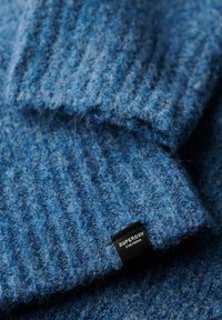 Tissu tricoté bleu avec une texture côtelée. Présente des fibres douces et duveteuses ainsi qu'une étiquette noire portant la marque "SUPERDRY" sur le bord.