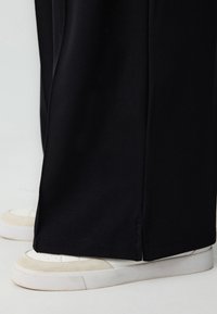 Pantalons larges noirs en tissu lisse ; dotés d'une subtile couture en bas. Associés à des baskets blanches avec des accents beiges.