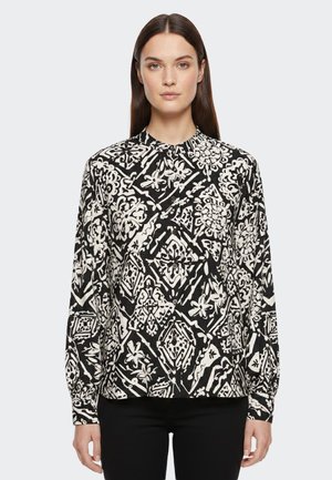 Donna con capelli lisci e scuri che indossa una camicetta a maniche lunghe con motivo bianco e nero e pantaloni neri, in piedi davanti a uno sfondo semplice.