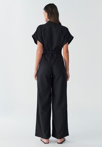 Juodas kombinezonas trumpomis rankovėmis, plačiomis kojų kišenėmis, su diržu juosmenyje ir matomomis siūlių detalėmis. Lygus audinys su matinio paviršiaus apdaila.