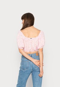 Top curto rosa com mangas bufantes e padrão floral; apresenta um decote quadrado nas costas, textura franzida e tecido suave. Combinado com jeans azuis.