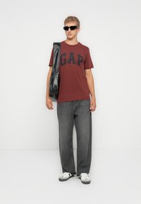 Bordeaux t-shirt med fed sort "GAP" skrift, parret med løstsiddende grå jeans og hvide sneakers. Mand bærer en sort tote bag.