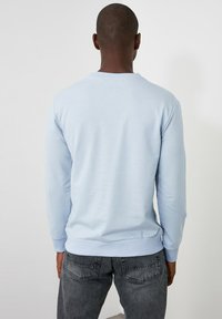 Sweatshirt bleu clair à manches longues avec poignets côtelés, doté d'un col rond. Le tissu présente une texture lisse; vue de dos montrant un design minimaliste.
