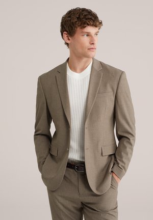 Noore mehe lokkis juustega, kes kannab helepruuni ülikonda ja valget kootud kampsunit, seisab käed taskutes ja vaatab küljele.