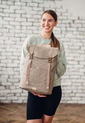 Brown fabric backpack with a flap closure, adjustable straps, and gold-tone hardware. Smooth texture, features a front pocket and decorative accents.