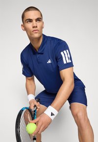 Námořnická modrá sportovní polo s bílými trojitými pruhy, spárovaná s odpovídajícími šortkami. Drží tenisovou raketu a tenisový míček, má na sobě zápěstní bandáže.