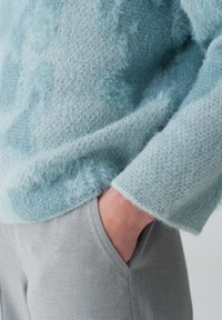 Strukturierter, hellblauer Strickpullover mit flauschiger Oberfläche, kombiniert mit grauen Hosen. Der Pullover hat eine lockere Passform und gerippte Bündchen.