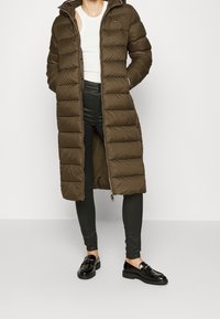 Giacca imbottita verde oliva con zip frontale e cappuccio, caratterizzata da sezioni trapuntate orizzontali. Indossata su leggings neri e mocassini.