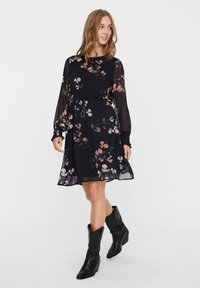 Robe noire à imprimé floral avec des manches longues, taille élastique et tissu transparent. Présente des motifs de fleurs roses et blanches. Associée à des bottes noires montantes.