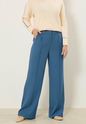 Kobieta ubrana w szerokie niebieskie spodnie, kremowy sweter z dzianiny z przeszyciami na rękawach oraz beżowe buty z zaostrzonym noskiem, stojąca na tle jednolitego tła.