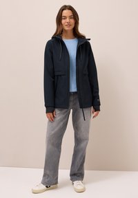 Mörkblå utomhusjacka med huva, dragkedja och framfickor. Bärs över en ljusblå topp och grå vida jeans tillsammans med vita sneakers.