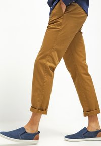 Homme marchant, portant un pantalon chino moutarde retroussé et des chaussures bleues à enfiler avec des semelles blanches sur fond blanc.