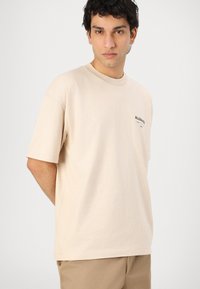Light beige cotton t-shirt, crew neck, short sleeves. Features small black "ALLSAINTS" logo on the left chest. Relaxed fit.