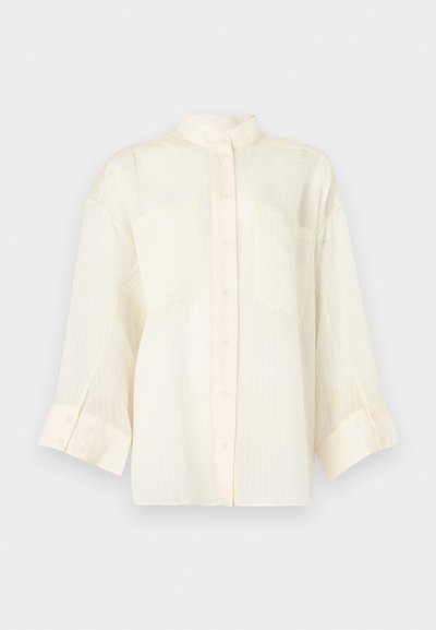 Blouse crème à manches longues avec boutons sur le devant, deux poches plaquées et un col montant, présentée sur un fond clair uni.