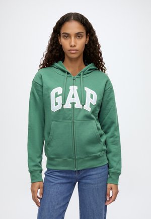 Une femme porte un sweat à capuche vert zippé avec l’inscription GAP en blanc et un jean bleu, sur un fond clair uni.