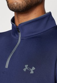 Tamno plavi pulover s četvrtastim zipom, izrađen od teksturiranog materijala, sivi zip i logo Under Armour na prsima.