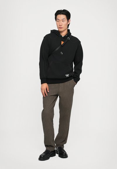 Black hoodie with contrasting seams, front pocket, and logo detail, paired with brown striped trousers and black shoes.
