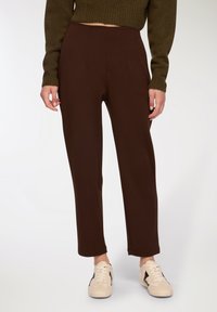 Brązowe zwężane spodnie o gładkiej fakturze, z przodu z zakładkami i średnim stanem w talii. W połączeniu z jasnymi trampkami.