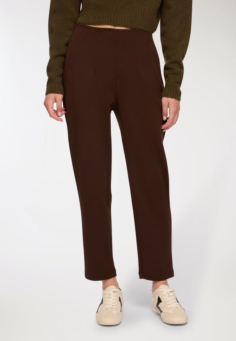 Brązowe zwężane spodnie o gładkiej fakturze, z przodu z zakładkami i średnim stanem w talii. W połączeniu z jasnymi trampkami.