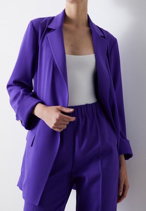 Frau trägt ein lila Blazer und passende hochgeschnittene Hose mit weißem ärmellosem Oberteil vor einem schlichten hellen Hintergrund.