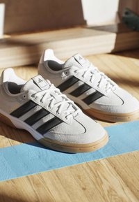 Weiße Adidas-Sneaker mit schwarzen Streifen, Gummisohlen und Schnürsenkeln auf einem Holzboden mit einem blauen Streifen.