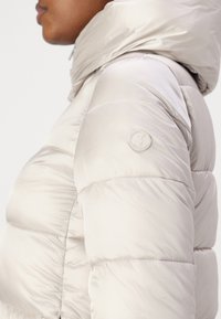 Svijetlo bež puffer jakna s visokim ovratnikom, kvadratnim dizajnom i okruglim logom na ramenu. Glatka, sjajna tekstura tkanine.