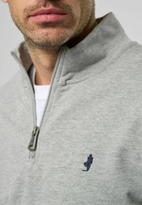 Grå genser laget av mykt stoff, med glidelås krage og en liten marineblå brodert logo på brystet. Glatt tekstur.