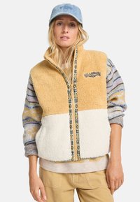 Fleece-jersey liivi, jossa on vaaleanruskea yläosa, kerma-alaosa ja kuvioitu vetoketju. Päällä monivärinen neulepitkähiha ja asusteena vaaleansiniset sortit.