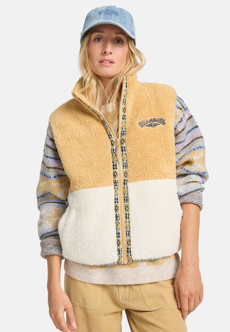 Fleece-jersey liivi, jossa on vaaleanruskea yläosa, kerma-alaosa ja kuvioitu vetoketju. Päällä monivärinen neulepitkähiha ja asusteena vaaleansiniset sortit.